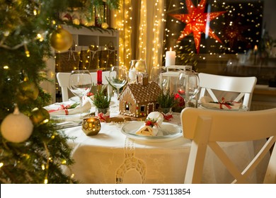 Beautiful served table with decorations, candles and lanterns. Little gingerbread house with glaze on white tablecloth. Living room decorated with lights and Christmas tree. Holiday setting