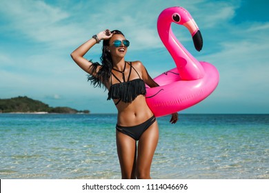 A beautiful sexy amazing young woman on the beach sits on an inflatable pink flamingo and laughs, has a great time, tanned perfect body, long hair, black bikini, fashion accessories, low key photo