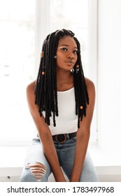 Beautiful sexy American black woman with stylish dreadlocks in fashion denim clothes near window in room on bright day. Fashion model black girl relaxes indoors. Youth style. Casual look.