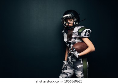 Beautiful sexy athletic woman poses on black background in American football uniform and helmet with ball in her hand. Copy space.
