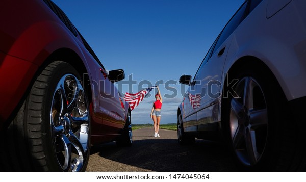 Beautiful sexy blonde girl with a sports figure\
gives the go-ahead to cars with the American flag, attention to the\
start. Ready, set, go. Sports cars at the start, speed and\
excitement. Top gear,