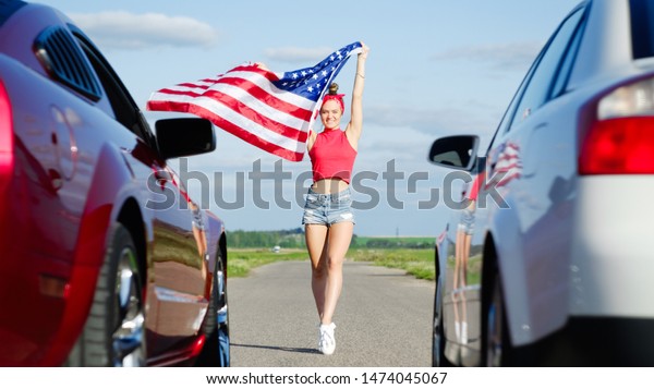 Beautiful sexy blonde girl with a sports figure\
gives the go-ahead to cars with the American flag, attention to the\
start. Ready, set, go. Sports cars at the start, speed and\
excitement. Top gear,