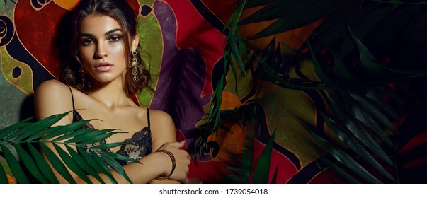 Beautiful sexy dark-skinned young girl posing against the backdrop of a motley colorful wall surrounded by palm leaves.