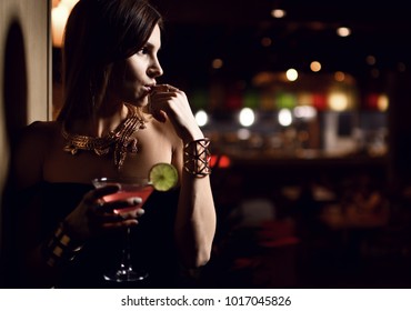 Beautiful sexy fashion brunette woman in expensive interior restaurant drink martini cosmopolitan cocktail on dark cafe background
