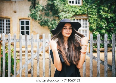 Beautiful sexy girl brunette with brown eyes in a black dress and a black hat with big brims against the backdrop of a wooden fence and an old house in a village, Burgundy in france in summer.