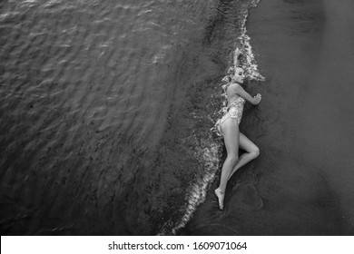 Beautiful sexy girl model lies on white sand beach, in a black swimsuit, with a tanned body, long blonde hair, magnificent breasts. Healthy nature, ocean waves. A young woman is lying on the sand 