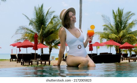 Beautiful sexy girl in white swimwear smiles enjoy relax with orange cocktail drink in resort. Young woman in straw hat sit on edge of outdoor swimming pool in luxury hotel. Tropical summer vacation.