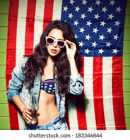 beautiful sexy long haired girl with glass bottle of water and skateboard against american flag sets sunglasses