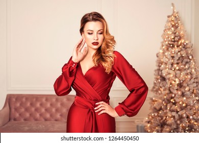 Beautiful Sexy Plus Size Model Woman In Glamour Red Dress, Christmas Tree On A Background. Curly Brunette Hair And Red Lips. Copy Space