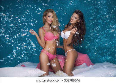 Beautiful Sexy Two Girls Having Fun In Bedroom, Pillow Fight. Blonde Woman And Brunette Lady Smiling, Looking At Camera. 