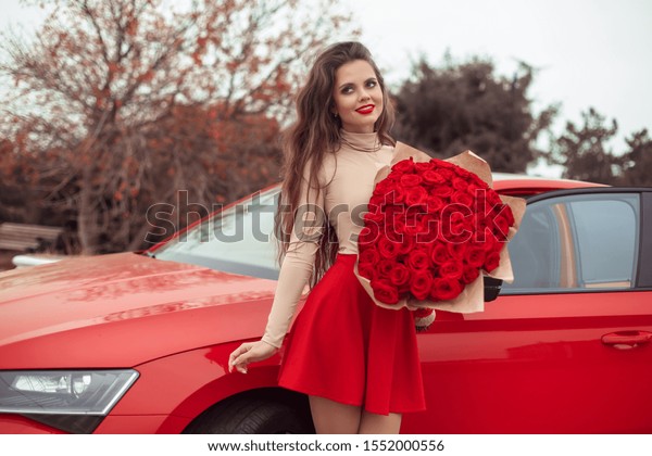 Beautiful sexy woman with rose bouquet of flowers posing\
by new red car. Charming brunette in red skirt. Beauty outdoor\
autumn portrait. Valentines day. Romantic expensive luxury gift.\
