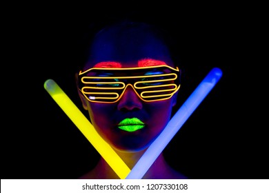 Beautiful sexy woman with UV face paint, glowing clothing, glowing bracelet in front of camera, face shot, confident look, holding light sticks. Asian woman. Party concept.