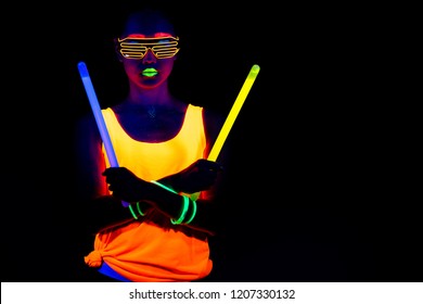 Beautiful Sexy Woman With UV Face Paint, Glowing Clothing, Glowing Bracelet In Front Of Camera, Half Body Shot, Confident Look, Holding Light Sticks. Asian Woman. Party Concept.