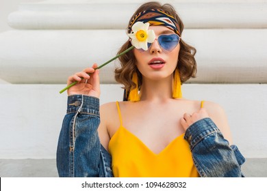 beautiful sexy woman in yellow stylish dress wearing denim jacket, holding flower, trendy outfit, hippie indie style, spring summer fashion trend, blue sunglasses, street fashion, trendy accessories