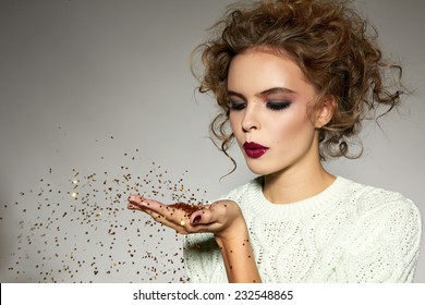 Beautiful sexy young blond girl with wavy hair bright evening make-up red lips long fluffy eyelashes holds palm with gold sequins in front of her and blows them a holiday New Year Christmas joy fun