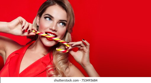 Beautiful sexy young girl in a red dress and long blond hair biting a festive candy cane on a red background.