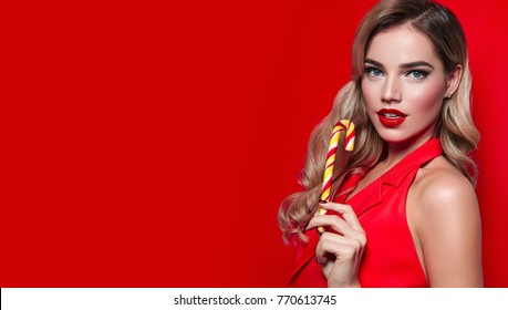 Beautiful sexy young girl in a red dress and long blond hair biting a festive candy cane on a red background.Fashion, beauty, make-up, cosmetics, hairstyle, beauty salon, boutique, discounts, sales.
