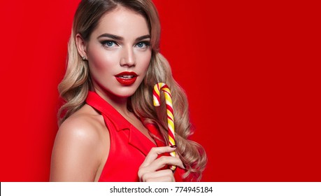 Beautiful sexy young girl in red dress and long blond hair is holding a festive candy cane in hand on a red background.