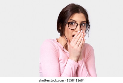 Beautiful Shocked Young Woman Wears Pink Long Sleeve Blouse, Looking At The Camera, With Hand On Mouth, Posing Over White Studio Background. Close Up Portrait Of Surprised Female  Feels Amazed.