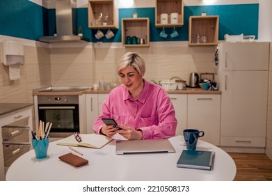 Beautiful Short Haired Blonde Woman Sits At The Kitchen Table And Looking In The Smartphone. Check Electronic Day Planner In Phone App And Planning A Meeting. Reading Email On A Cell Phone.