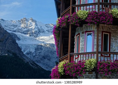 Beautiful Shot Of Colorful Flowers Covering The Wooden Balcony Of A Stone Hotel High In The Tranquil Italian Mountains. Picturesque View Of The Snowy Mountain Range Behind A Lovely Rustic Hotel.