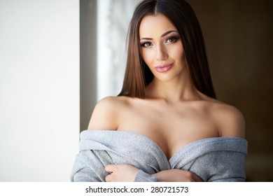 Beautiful showy girl with plump lips and big Boobs in gray soft sweater with bare shoulders and bare back standing near the window. Sexy, shy lady