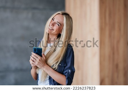 Beautiful shy blonde laughing swedish girl in grey shirt holding phone toothy smiling indoors. Attractive Caucasian young woman enjoying new cellphone. Electronics and gadgets.