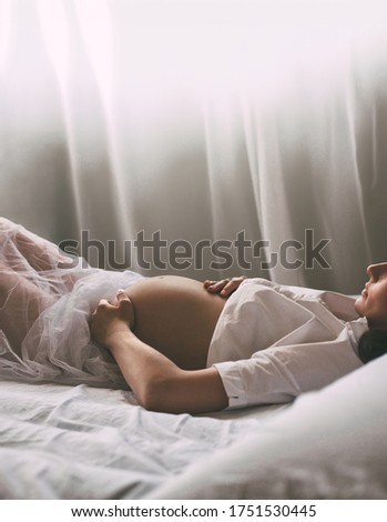 Beautiful silhouette of a pregnant woman with a bare stomach. A female in a white shirt lies on the bed and touches her stomach