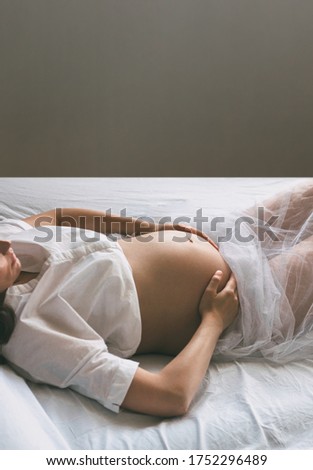 Beautiful silhouette of a pregnant woman with a bare stomach. A female in a white shirt lies on the bed and touches her stomach