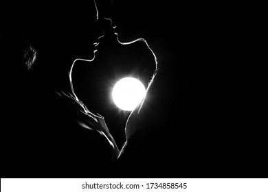 Beautiful silhouettes of a man and a woman are facing each other, between a mini silhouette of a heart and the bright light of a distant moon. The couple’s first kiss is in love. First love forever
