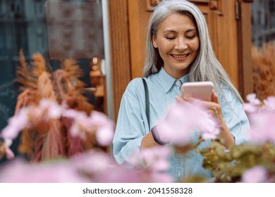 Beautiful silver haired mature Asian woman in denim shirt uses mobile phone on outdoors terrace decorated with flowers