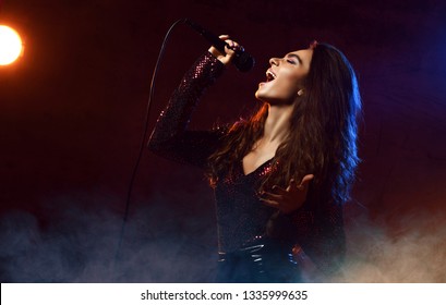Beautiful singing girl curly afro hair. Beauty woman singer sing with microphone karaoke song on stage. Dark background, smoke, spotlights