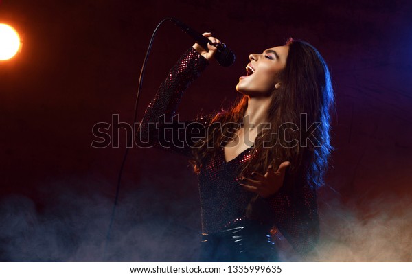 Beautiful singing girl curly afro hair. Beauty\
woman singer sing with microphone karaoke song on stage. Dark\
background, smoke,\
spotlights