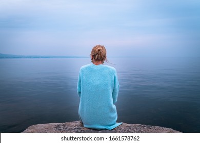 Beautiful Single Solo Young Traveler Blond Woman With Tranquit Jacket Sitting Alone On Stone Looking In To Water In The Blue Ocean Foggy Mist Lake In Cold Winter With Sad Dark Depression  Emotional