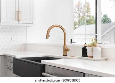 A beautiful sink in a remodeled modern farmhouse kitchen with a gold faucet, black farmhouse sink, white granite, and a tiled backsplash. No labels.