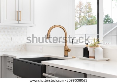 A beautiful sink in a remodeled modern farmhouse kitchen with a gold faucet, black farmhouse sink, white granite, and a tiled backsplash. No labels.