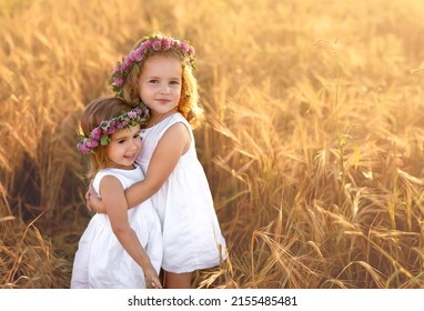 Beautiful sister girls in white dresses with wreaths of purple clover flowers on their heads are hugging in a field with ears of wheat. Best friends since childhood and forever. Strong friendship 