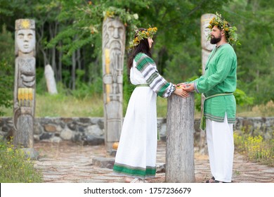 Beautiful slavonic couple with flower crowns in white and green embroidered ethnic costumes holding hands against the background of a forest and slavic idols on a summer day. Holiday of Ivan Kupala.