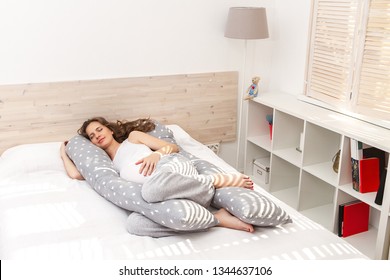 Beautiful sleeping smiling pregnant woman with a big tummy lying in an anatomical comfortable ergonomic body pillow on the bed indoor.