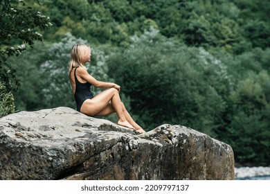 A Beautiful Slender Blonde Girl In A Dark Joint Swimsuit Sits On A Large Stone Near A Mountain River. Rest In Exotic Places Of Nature