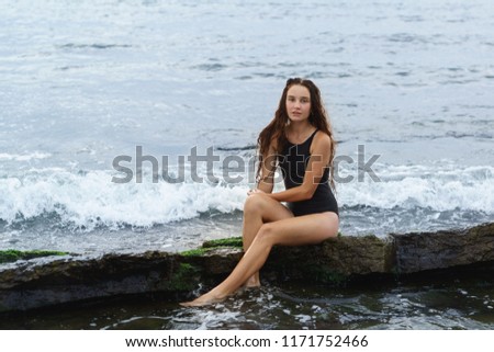 A beautiful slender long-haired girl in a black swimsuit sits on a stone on the sea. Cloudy summer day