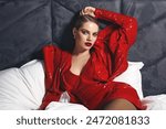 A beautiful slender young luxurious woman in a short red shiny dress lies on white pastel linen in the bed. Advertising fashionable clothes. Advertising of decorative cosmetics. 