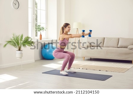 Beautiful and slender young woman performs sports exercises at home using dumbbells. Smiling girl in comfortable sportswear sitting down slightly and stretching her arms forward holding two dumbbells.