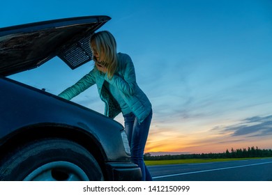 Beautiful Slim Blonde Girl Talking On The Phone And Looking Under The Hood Of A Car. The Girl Will Break Down The Car In The Evening At Sunset, Need Help.