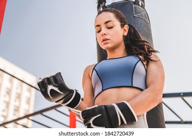 Beautiful Slim Female Sportsman Standing With Punching Bag Behind Wearing Boxig Gloves Outside