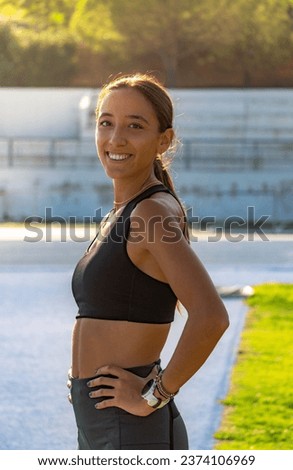 A beautiful slim girl dressed in tight sports clothing, looking and smiling happily towards the camera and hands on her waist illuminated from behind by the evening sunlight.