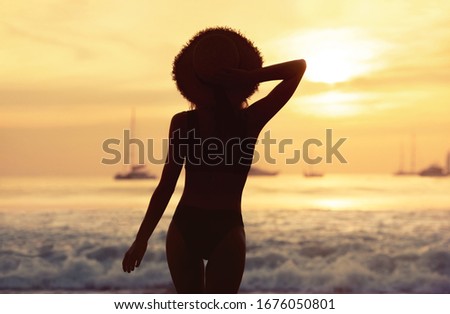 Beautiful slim girl in swimsuit and hat stands at sunset sea beach