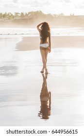 Beautiful slim tanned woman with long black hair walking alone on paradise beach in Bali at sunset. Nobody around and mirror reflection in calm water surface. Idyllic and dreamlike view