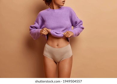 Beautiful slim women body. Photo of fit woman takes off purple sweater shows her flat belly wears panties has healthy skin isolated over beige studio wall
