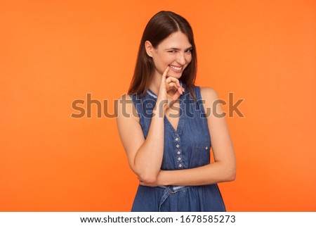Beautiful and sly brunette woman in denim dress pondering mysterious tricky plan, smiling with cunning playful expression, scheming evil plan, devious idea. studio shot isolated on orange background